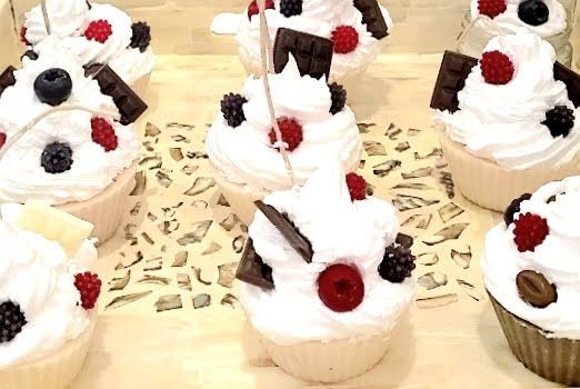 Cupcakes topped with whipped cream, berries, and chocolate pieces on a decorative surface.